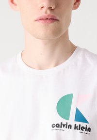 Balta medvilninė marškinėlių palaidinė su teal ir rožinės spalvos grafika, po logotipu esantiame tekste "calvin klein" ir "West 39th Street". Paprasta apvali iškirptė.