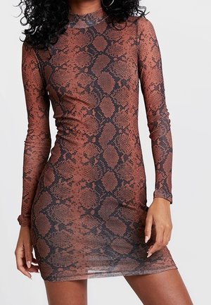 Kobieta ubrana w dopasowaną, długorękawową sukienkę w brązowo-czarny wzór wężowej skóry, z kolczykami-kołami, stojąca na tle jednolitego białego tła.