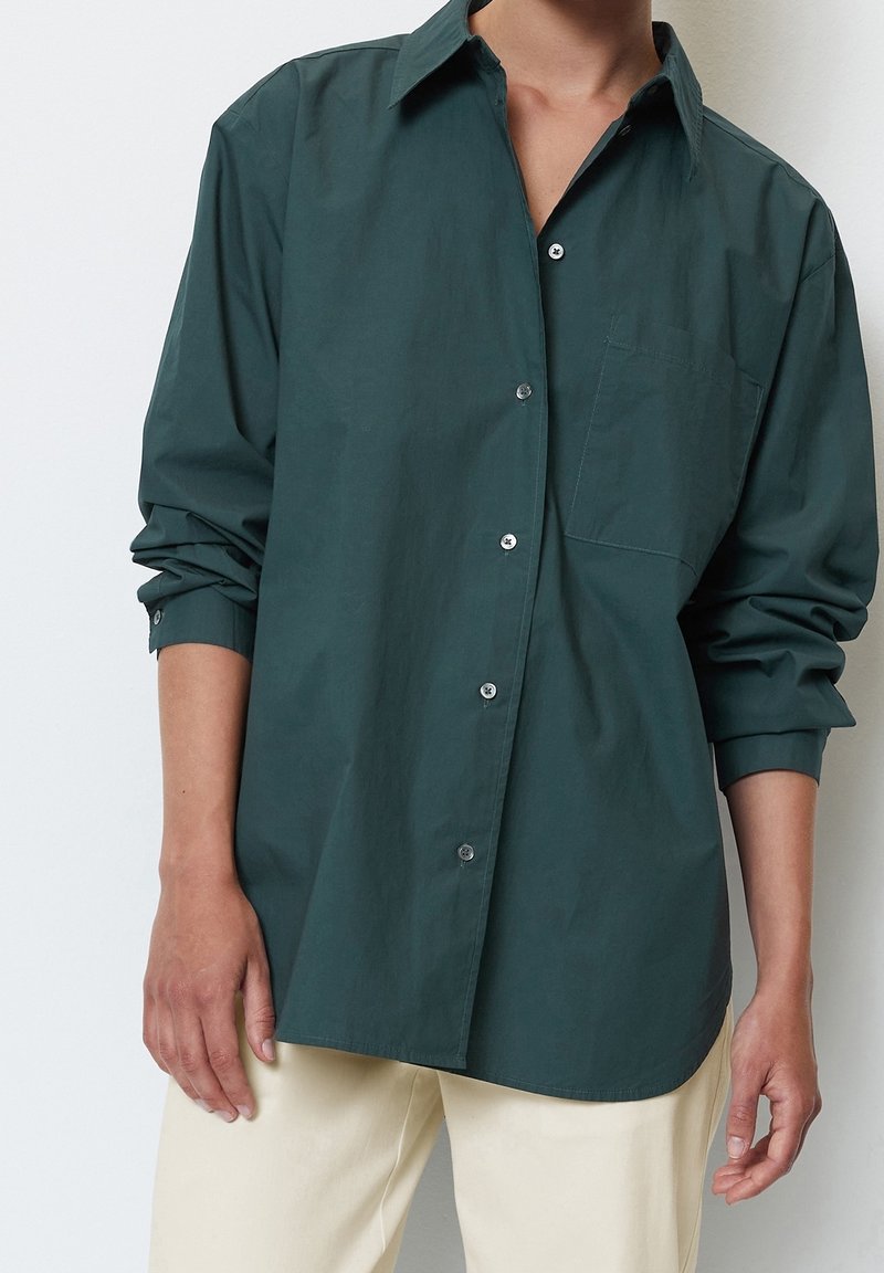 Chemise boutonnée vert foncé avec col classique, manches longues et une poche poitrine. Tissu lisse et léger avec des boutons en argent.