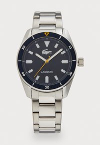 Montre en acier inoxydable avec un cadran rond bleu marine, des index horaires blancs et des accents jaunes. Équipée d'un bracelet en maillons argentés et d'une couronne pour les réglages.