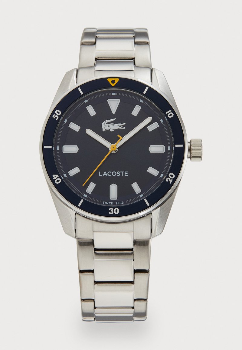 Montre en acier inoxydable avec un cadran rond bleu marine, des index horaires blancs et des accents jaunes. Équipée d'un bracelet en maillons argentés et d'une couronne pour les réglages.