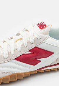 Vita och beige sneakers med röd läderlogotyp, vita snören och gummisulor, visar ovansidan och tungan med röd branding.