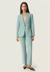 Világoskék, szabott öltöny mély V-nyakú blézert és egyenes szabású nadrágot tartalmaz. Az anyag sima tapintású. Fekete díszítésekkel készült.