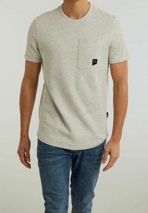 Mand iført en lysegrå, kortærmet tekstureret T-shirt med en brystlomme og blå jeans, stående mod en ensfarvet baggrund.