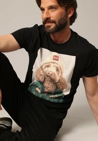 Zwarte t-shirt met een grafische print van een hond met een beanie en jack. Het ontwerp heeft zachte, realistische texturen en details.