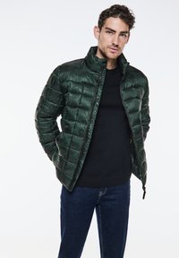 Opgevulde groene jas met een hoge kraag, voorzien van een quiltpatroon, een ritssluiting aan de voorkant en zijzakken, gecombineerd met een zwarte sweatshirt en blauwe spijkerbroek.