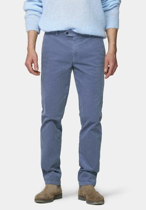 Mann trägt hellblauen Pullover, blaue Cordhosen und braune Wildlederstiefel, steht vor einem einfarbigen hellen Hintergrund.