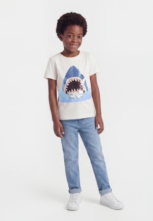 Smilende barn med kort krøllet hår iført en hvid t-shirt med en stor blå haj grafik, lyseblå jeans og hvide sneakers.