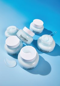 Viisi purkkia Torriden rauhoittavaa voidetta valkoisilla kansilla, joissa on vaaleansinistä voidetta, sinisen taustan edessä.