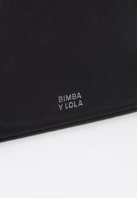 Melnas auduma soma ar gludu tekstūru un metāliski sudraba logo "BIMBA Y LOLA", kas izšūts priekšpusē.