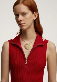 Top sin mangas de punto acanalado rojo con cremallera, combinado con un colgante circular dorado que presenta líneas texturizadas y en espiral, además de un detalle recortado.