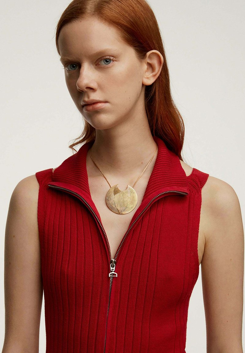 Top sin mangas de punto acanalado rojo con cremallera, combinado con un colgante circular dorado que presenta líneas texturizadas y en espiral, además de un detalle recortado.