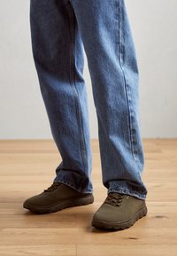 Zapatillas deportivas marrones con textura de malla y detalles de cordones, combinadas con unos jeans de mezclilla azul holgados, sobre un fondo de suelo de madera.