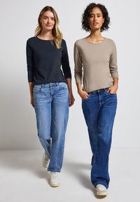Två individer bär långärmade tröjor; en marinblå och den andra ljusbeige. Båda har på sig ljusblå jeans och vita sneakers.