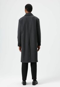 Lange donkergrijze jas van een wolmix, met een kraag met inkeping, rechte silhouet en twee zijnaden; gedragen over een zwarte broek en schoenen.