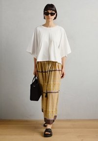 Weißes übergroßes Oberteil mit kurzen Ärmeln, gelbe gemusterte weite Hosen mit dunklen Streifen, schwarze Tasche und schwarze Sandalen. Glatte Texturen.