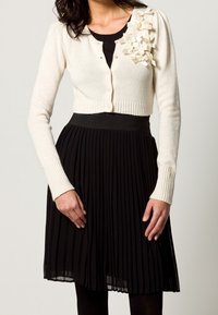 Krämfärgad kardiagan med ribbade fållar, prydd med texturerade vita tygdetaljer på vänster axel, tillsammans med en svart veckad kjol.