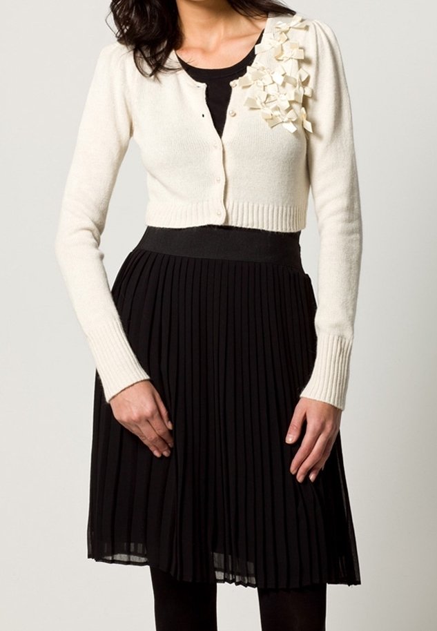 Krämfärgad kardiagan med ribbade fållar, prydd med texturerade vita tygdetaljer på vänster axel, tillsammans med en svart veckad kjol.