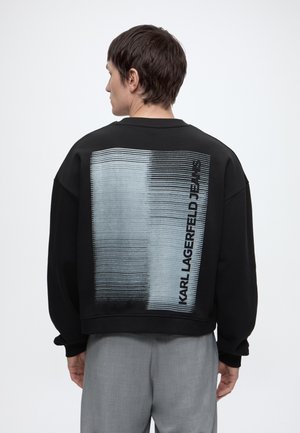 Osoba z krótkimi ciemnymi włosami nosi czarną bluzę z pionowymi liniami gradientowymi i napisem „KARL LAGERFELD JEANS” na plecach.
