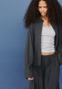 Jeune femme aux cheveux bouclés regardant vers le bas, portant un débardeur gris sous une chemise noire oversize et un pantalon noir assorti, le tout sur fond bleu.