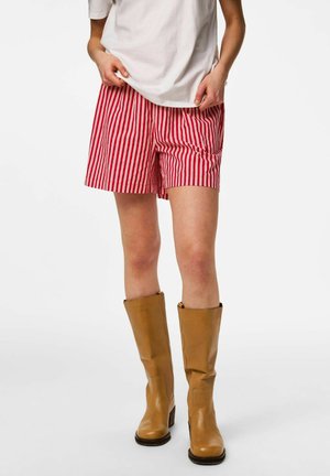 Persoon draagt rood-wit verticaal gestreepte short, een wit hemd en halflange lichtbruine leren laarzen, staand voor een effen achtergrond.