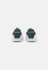 Un paio di sneakers bianche con linguette in suede verde scuro sul tallone, logo Lacoste visibile, fotografate da dietro su uno sfondo bianco.