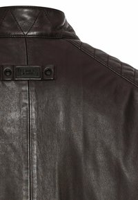 Brun læderjakke med et kviltet design på kraven og skuldrene, samt et mærket sort hardware-tag bagpå.