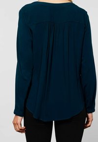 Blusa lunga a maniche con colore verde scuro, con un design sul retro a pieghe, scollatura a barca e orlo arrotondato. Tessuto liscio con polsini abbottonati.