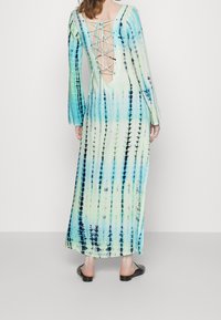 Dlouhé šaty s batikovaným vzorem v pastelových zelených a modrých odstínech, vykrojeným zadním dílem, šněrováním a dlouhými rukávy; látka má hladkou texturu.