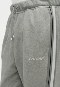 Grå joggingbukser med elastisk taljebånd og snore, sidelæns stribet detalje og "Calvin Klein"-logo broderet nær lommen.