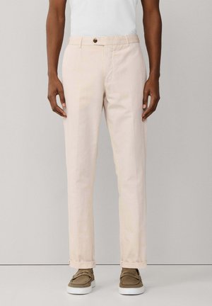 Persona vistiendo pantalones beige con puños, camiseta blanca y zapatos marrones sin cordones, de pie contra un fondo gris liso.