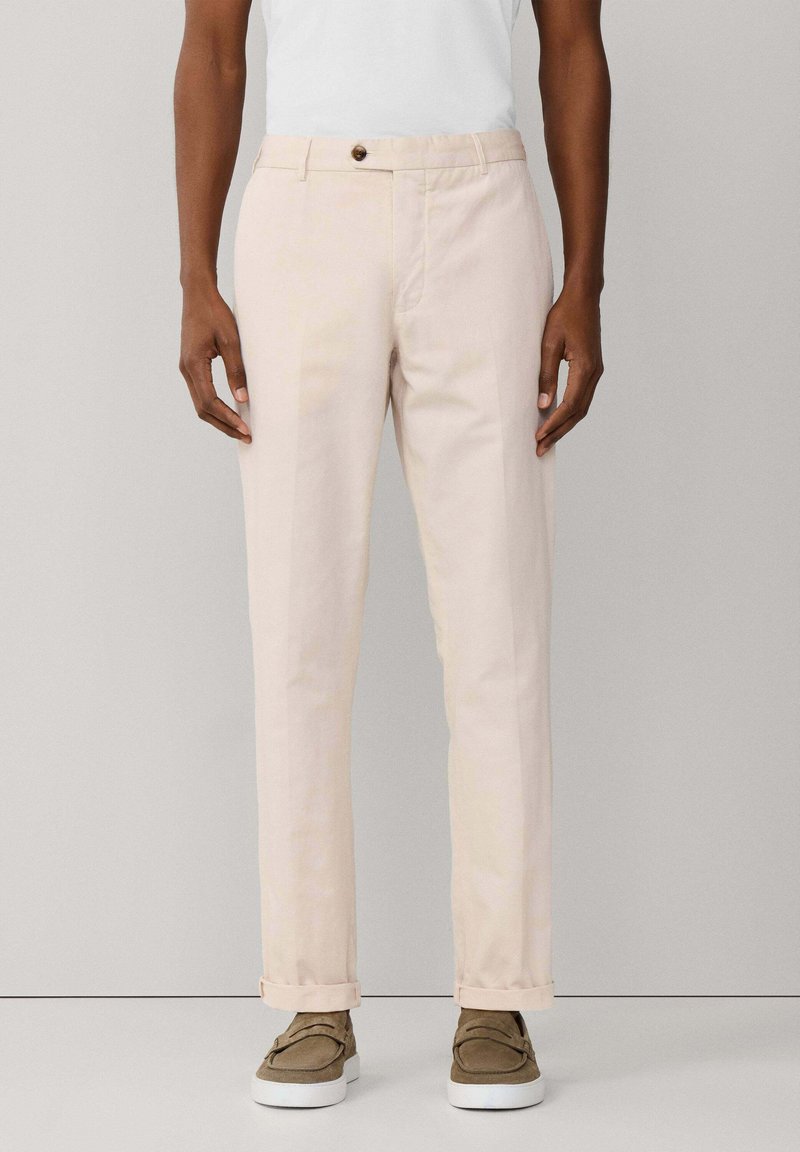 Persona con pantaloni beige risvoltati, maglietta bianca e scarpe marroni slip-on in piedi davanti a uno sfondo grigio uniforme.