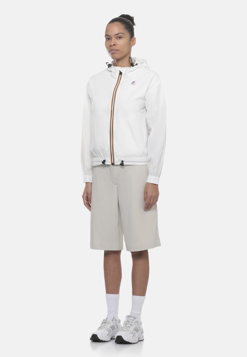 Žena stojaca, oblečená v bielej kapucňovej bunde s pásikavým zipsom, béžových kraťasoch po kolená, bielych ponožkách a bielych teniskách na bielom pozadí.