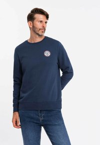 Sudadera azul marino hecha de tela suave, con cuello redondo, mangas largas y un logo de parche redondo en el pecho. Diseño casual con puños acanalados.