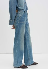 Jean large bleu clair en denim, avec une taille haute, des poches avant et un léger dégradé le long des jambes. Associé à des chaussures marron.