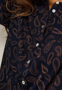Chemise bleu marine foncé avec un motif paisley complexe en brun clair, col à revers et boutons blancs. Tissu texturé.