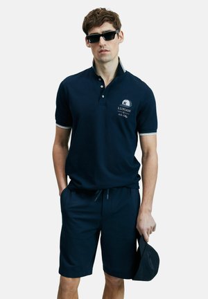 Man wearing navy polo shirt and shorts, black sunglasses, holding a navy cap in one hand, standing against a plain background.