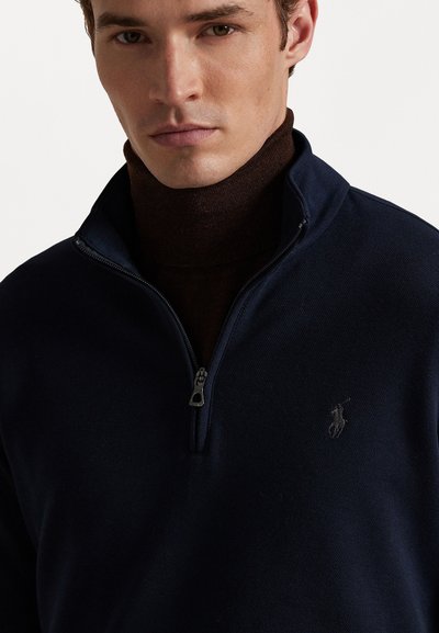 Marineblå hettegenser med glidelås og en teksturert overflate, samt en liten brodert logo. Har høy krage og en kontrasterende brun toroms under.