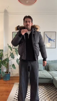 Svart puffjakke med hette fôr med pels, glidelås foran og sidelommer, brukt over en mørk genser og kombinert med løse svarte bukser.