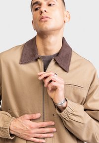 Beige canvas jacket with a brown corduroy collar, zipper closure, and elastic cuffs, featuring a chest pocket and subtle stitching details.