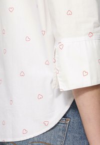 Chemise blanche avec des motifs de cœurs rouges dispersés, dotée d'un ourlet arrondi et de manches retroussées. Texture lisse, associée à un jean en denim.