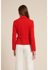 Czerwony wełniany blezer o dopasowanym kroju, z zarysowanymi ramionami i klapą z wycięciem. Posiada rozcięcie z tyłu oraz przeszycia nadające mu dopasowany wygląd.