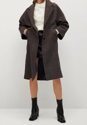 Femme portant un long manteau à carreaux marron, un pull blanc, une jupe en cuir noire et des bottines noires, debout devant un fond uni.