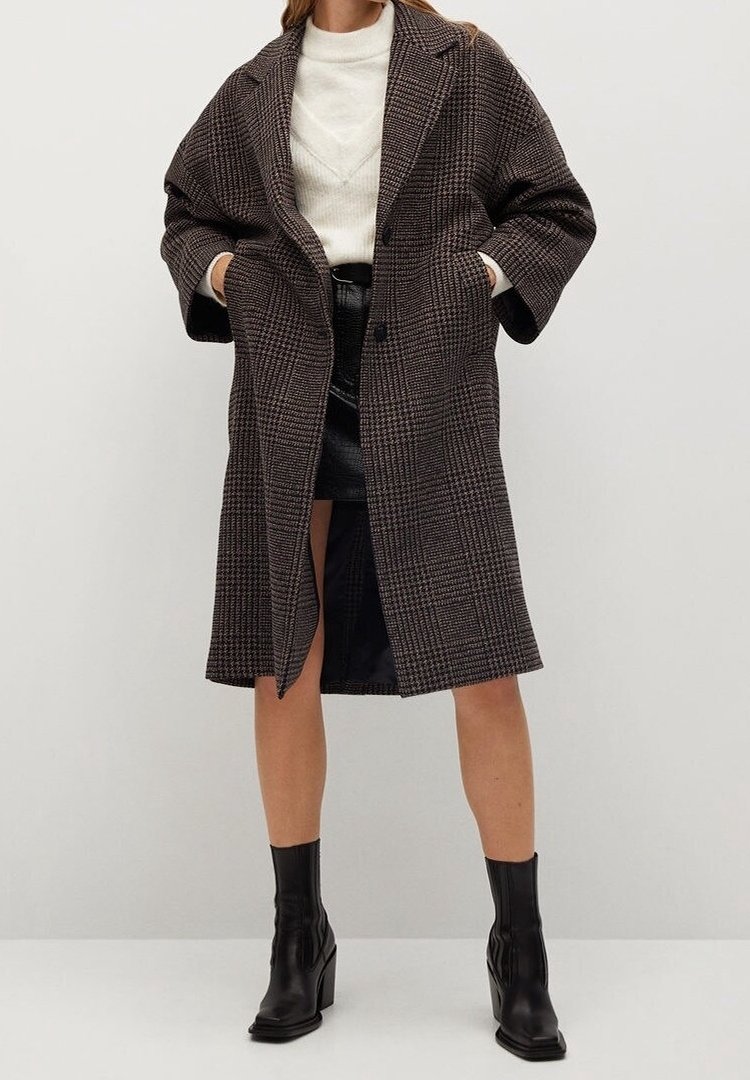 Femme portant un long manteau à carreaux marron, un pull blanc, une jupe en cuir noire et des bottines noires, debout devant un fond uni.