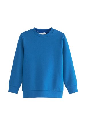 Kind mit lockigem Haar lächelt über die Schulter, trägt einen blauen Pullover, weißes Hemd mit Kragen und schwarze Hose vor einem einfachen Hintergrund.