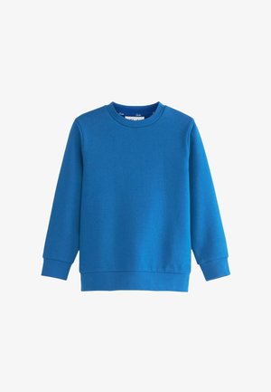 Enfant aux cheveux bouclés sourit par-dessus l'épaule, portant un sweat bleu, une chemise blanche à col et un pantalon noir sur un fond uni.