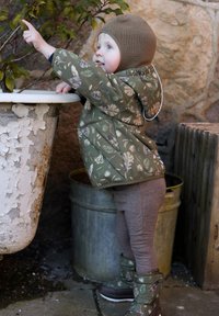 Bambino con giacca trapuntata verde oliva con motivi floreali, leggings grigi e stivali decorati, che si protende verso una pianta accanto a un vaso consumato dal tempo.