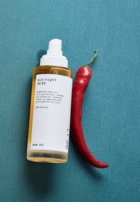 Ambra glassflaske med kroppsolje med hvit pumpe, merket "mid/night 00.00," ved siden av en rød chilipepper på en blågrønn bakgrunn.