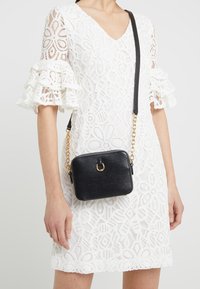 Bolso cruzado de cuero negro con una superficie texturada, herrajes dorados y una correa de cadena. Diseño compacto cuadrado con un detalle decorativo de anillo.