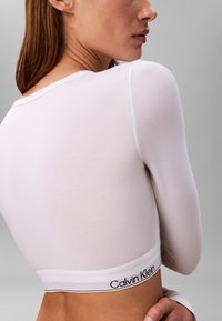 Maglietta bianca a maniche lunghe realizzata in tessuto liscio. Presenta un collo rotondo e una banda con logo sul fondo. Design semplice e aderente.